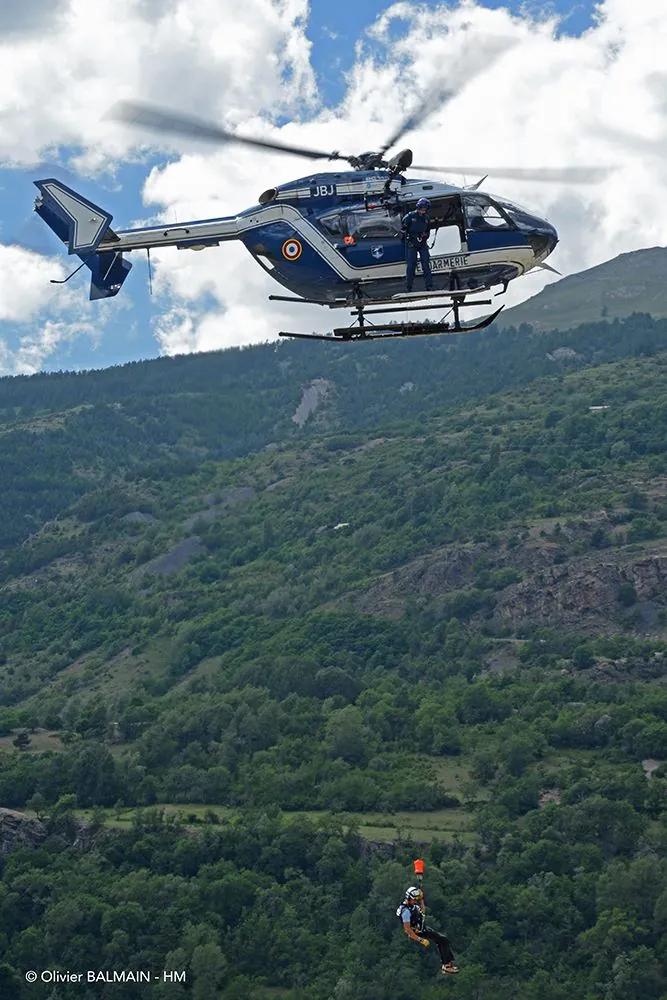 Hélicoptère EC145 bleu de la Gendarmerie en vol stationnaire, un sauveteur suspendu au câble du treuil