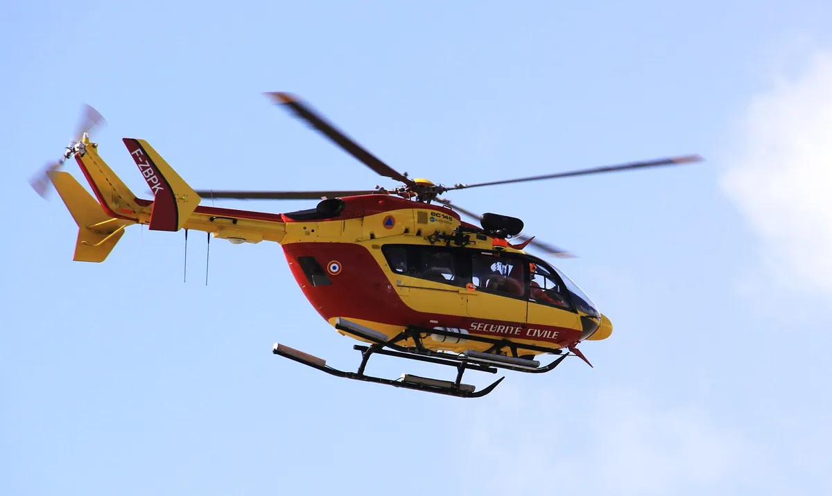 Hélicoptère EC145 rouge et jaune de la Sécurité civile survolant des mâts de voiliers dans un port