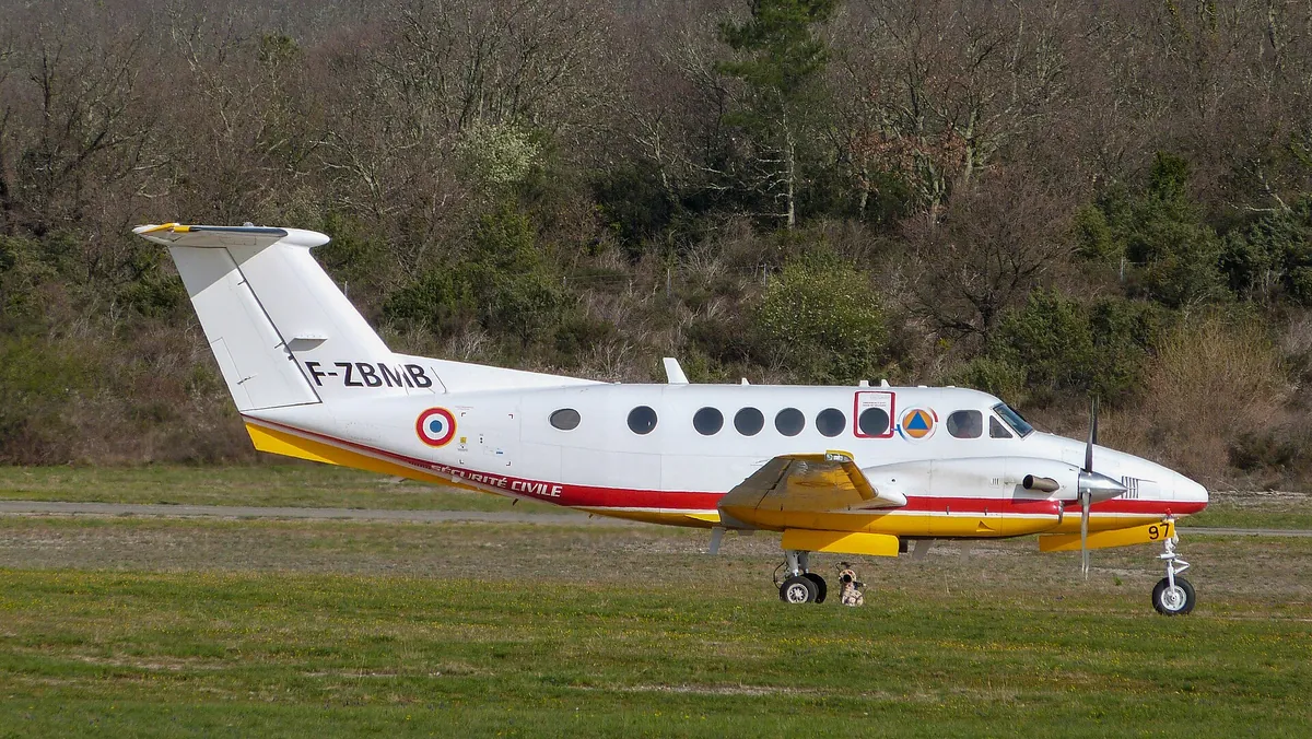Bimoteur Beechcraft King Air blanc à liseré jaune et rouge, immatriculé F-ZBMB, roulant sur une piste en herbe