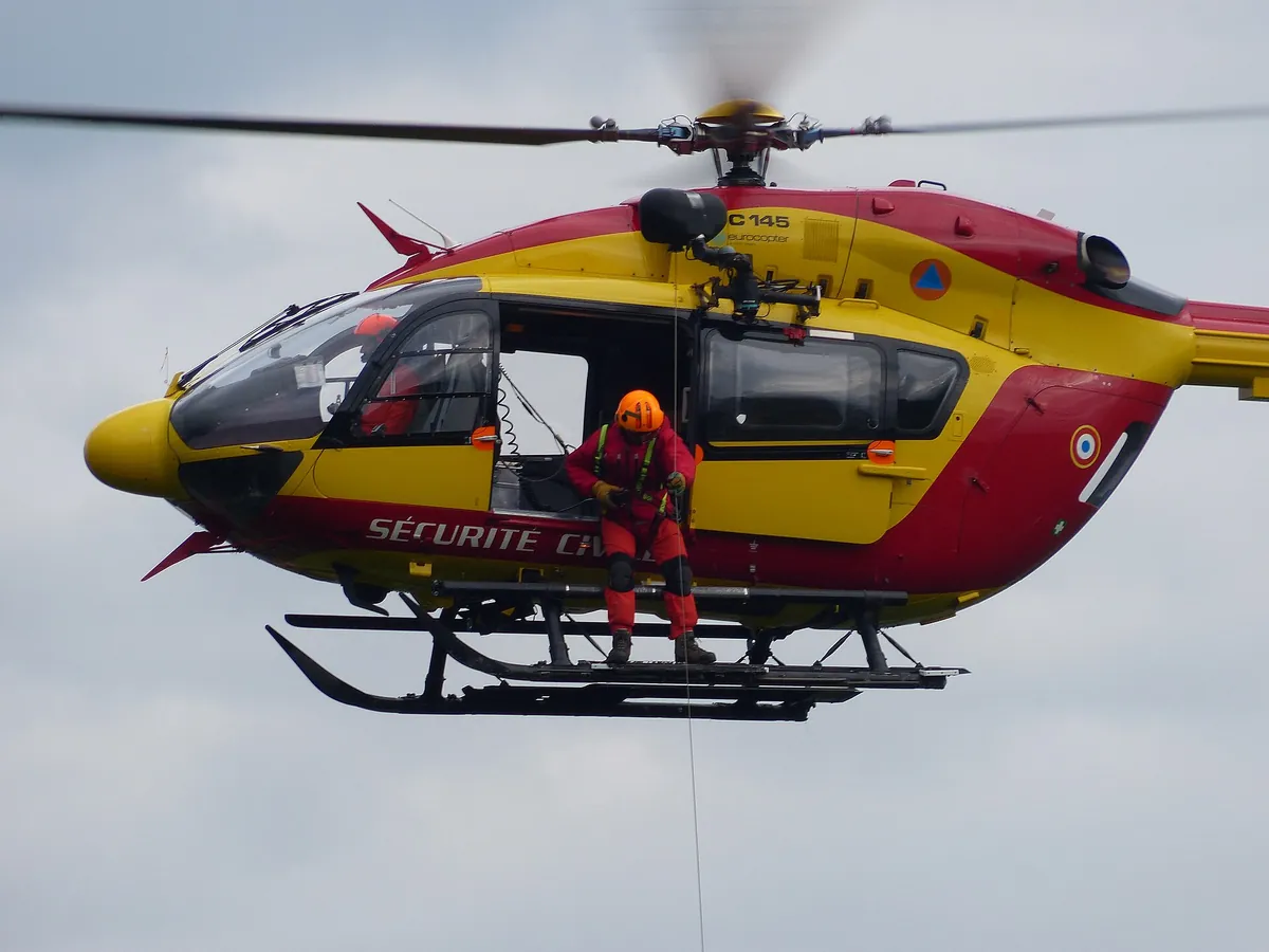 Sauveteur en combinaison orange suspendu au treuil sous un hélicoptère jaune et rouge de la Sécurité civile